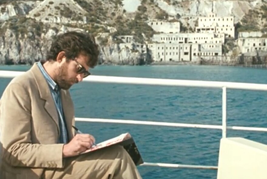 A man seated on a boat, writing in a book, with white buildings on a cliff against a blue sea.