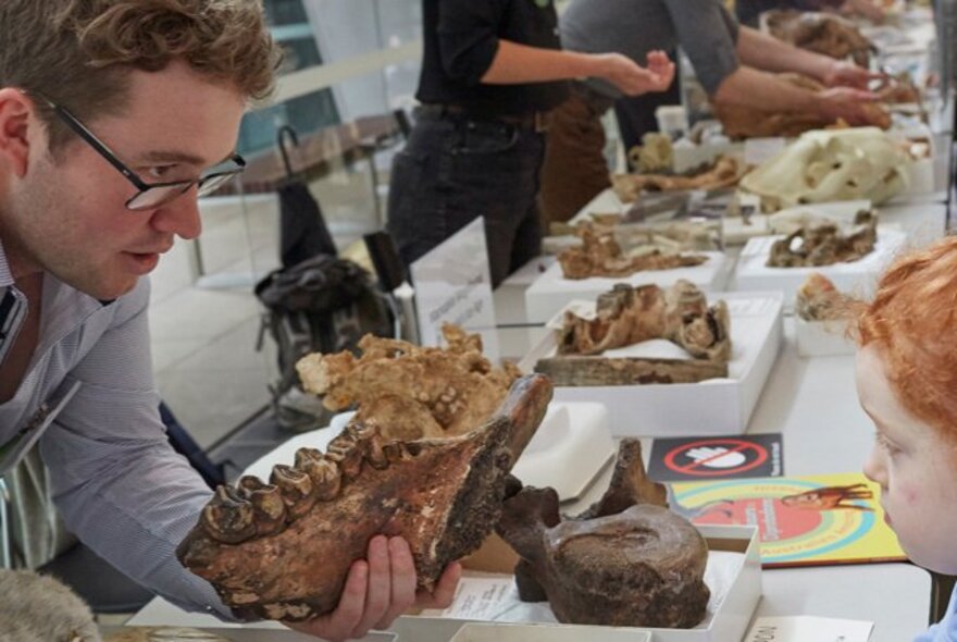A children speaking to an adult holding a large fossil, at a table displaying museum exhibits.