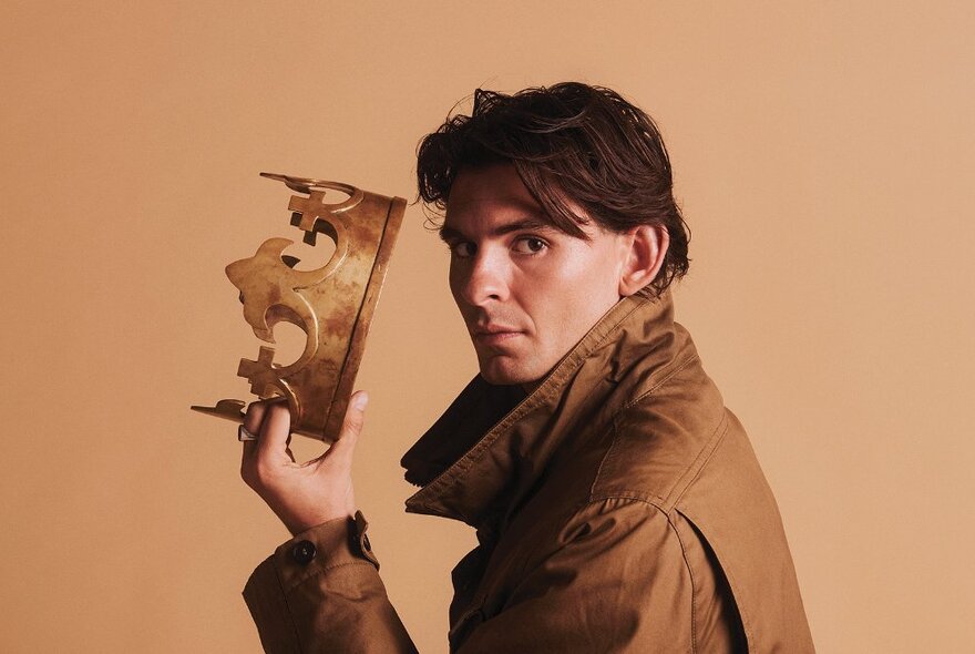 Side on view of a man in a brown raincoat with the collar turned up holding a crown up to his head, looking to his sharp left directly at the camera.