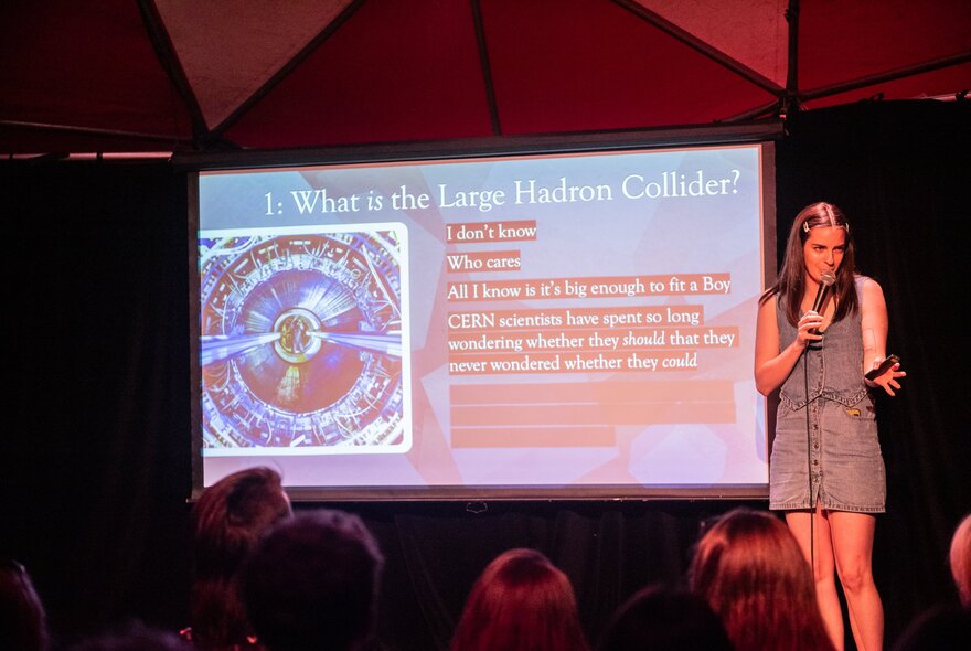 A performer on stage, talking into a microphone she holds, an audience in front of her, behind her is a screen with a slide presentation on it