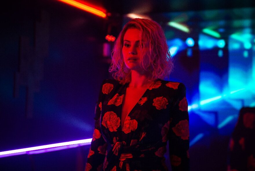 A still from the movie Emilia Pérez with a woman in a floral printed blazer in a dark area with neon lights behind her.