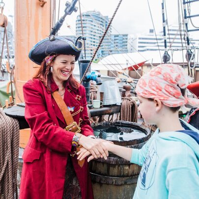 A person dressed as a pirate in a red coat shaking hands with a child wearing a head scarf aboard an old sailing ship.
