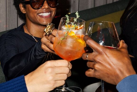 A person holding an orange cocktail, likely an Aperol Spritz, while toasting with friends.