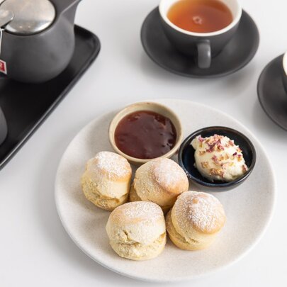 Plate of scones with cream and jam, accompanied by two cups of tea and a black teapot on a white surface.