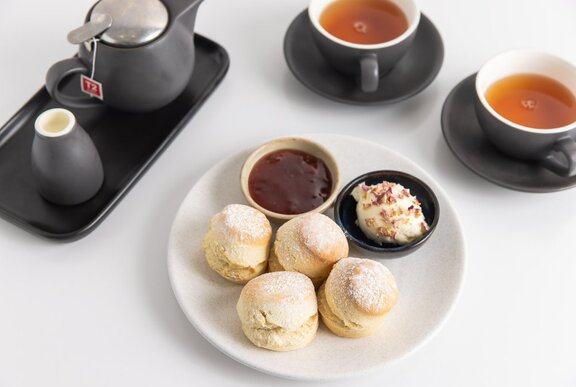 Plate of scones with cream and jam, accompanied by two cups of tea and a black teapot on a white surface.