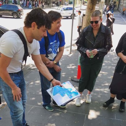 Tour leader showing three walking tour participants a page from an open notebook he holds, on a Melbourne city footpath.