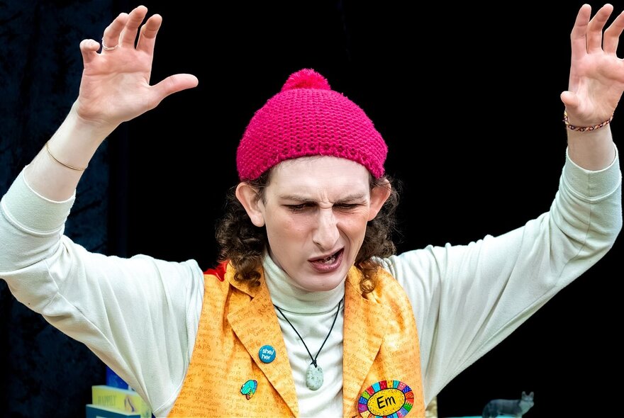 Person wearing red hat, yellow waistcoat and green skivvy, with hands in the air, pulling a twisted face.