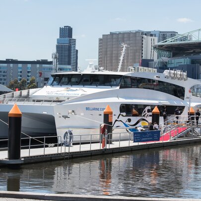 The Bellarine Express catamaran docked at the Docklands Terminal on Harbour Esplanade in Melbourne, with water in the foreground and city buildings in the background.