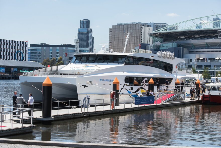The Bellarine Express catamaran docked at the Docklands Terminal on Harbour Esplanade in Melbourne, with water in the foreground and city buildings in the background.