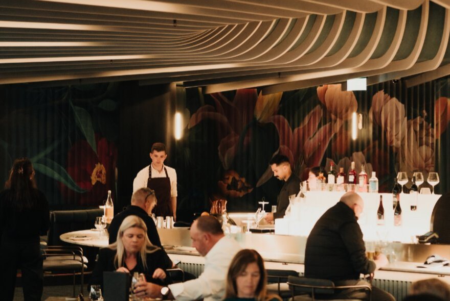 People seated in Amphora with wait staff looking on and a bartender at work. 