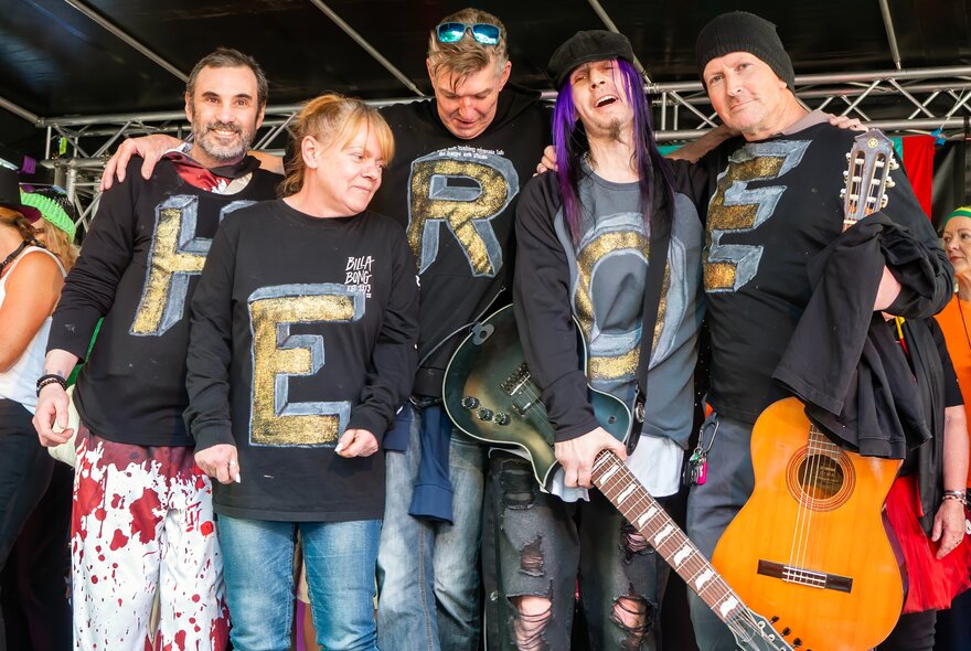 Five individuals standing on a stage wearing matching black long-sleeve shirts that together spell out the word H-E-R-O-E, two of them holding guitars.