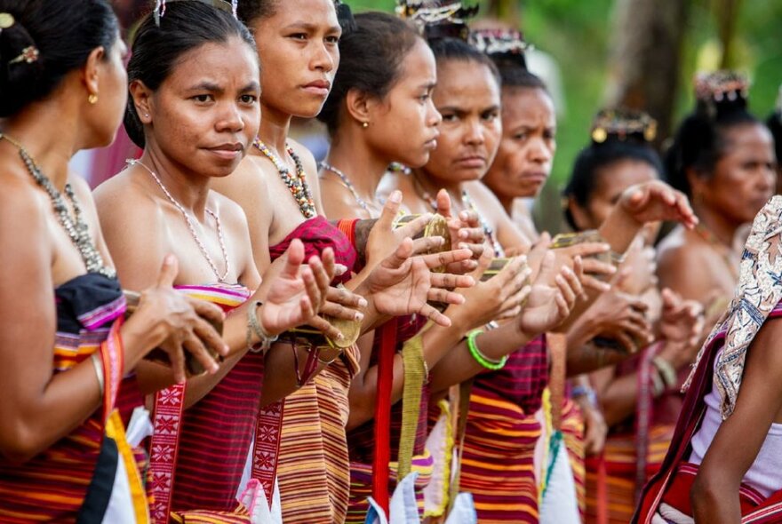 A row of women standing outdoors in Timor-Leste wearing handwoven textiles, their hands moving in front of them as if clapping. Photo: Jorge de Araujo