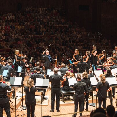 An orchestra performs while standing on a stage in front of a large, packed audience in a concert hall.