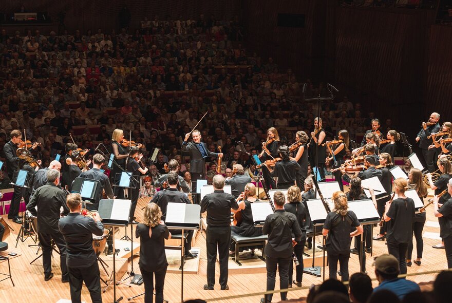 An orchestra performs while standing on a stage in front of a large, packed audience in a concert hall.