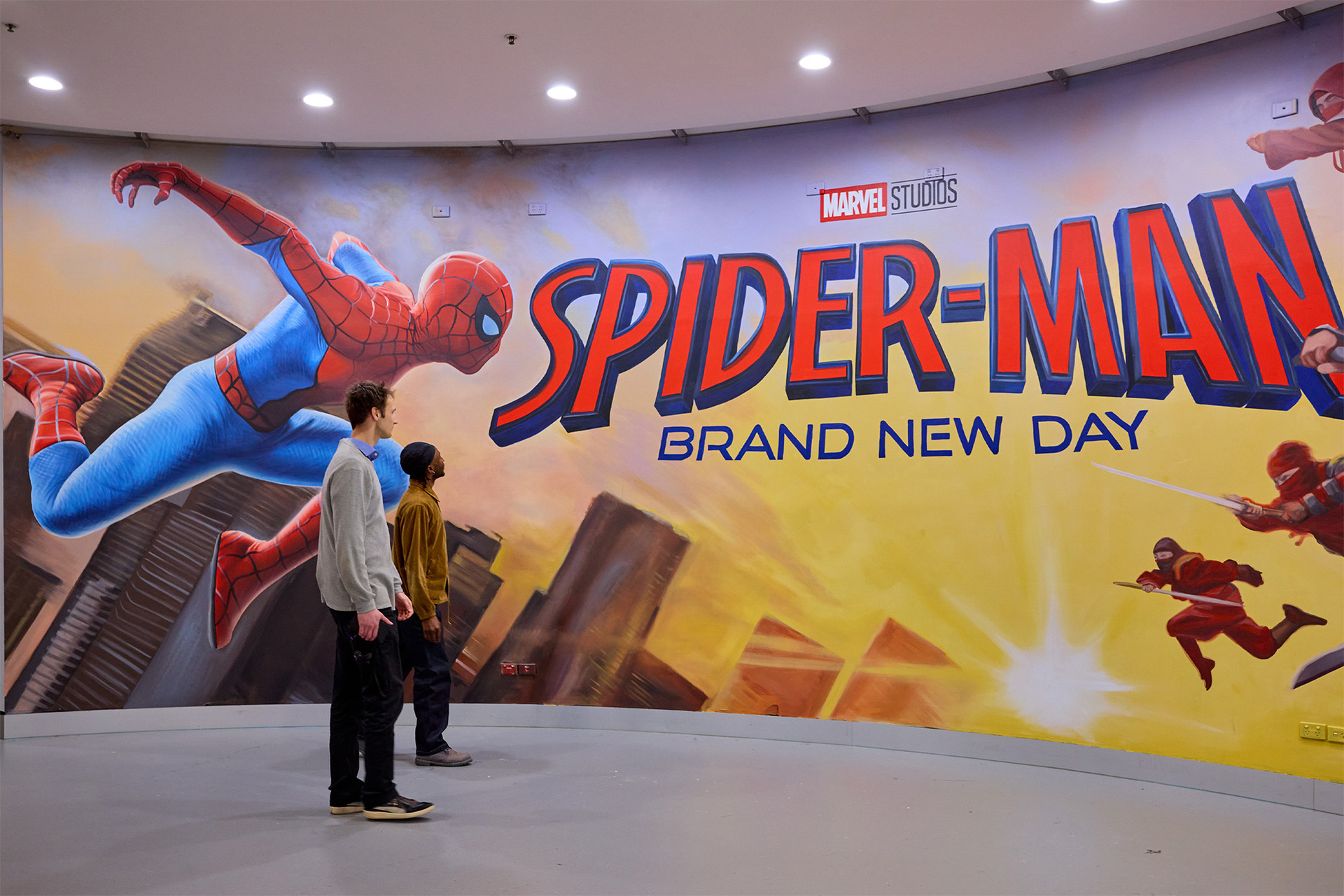 Two people are walking past a giant spider man poster