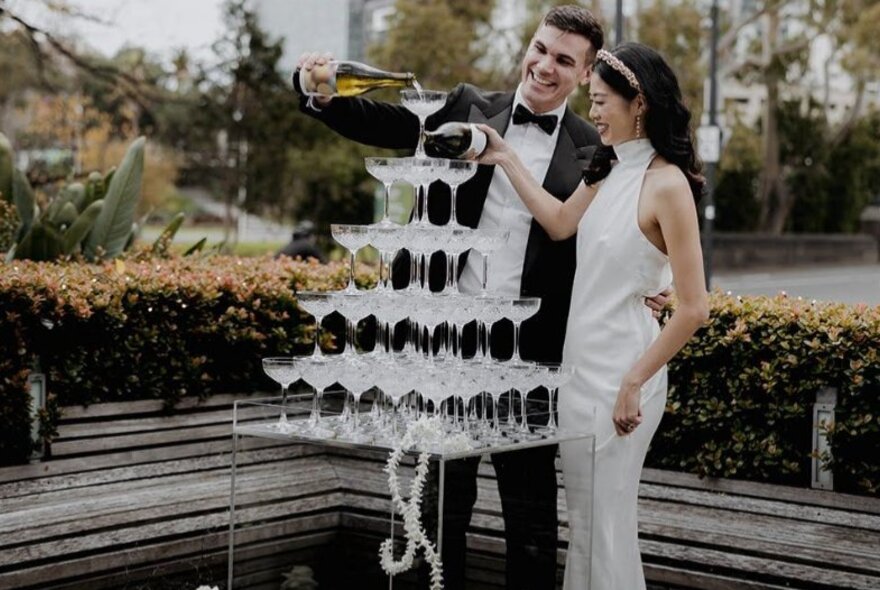 A wedding couple pouring bubbles into tiered rows of glasses.