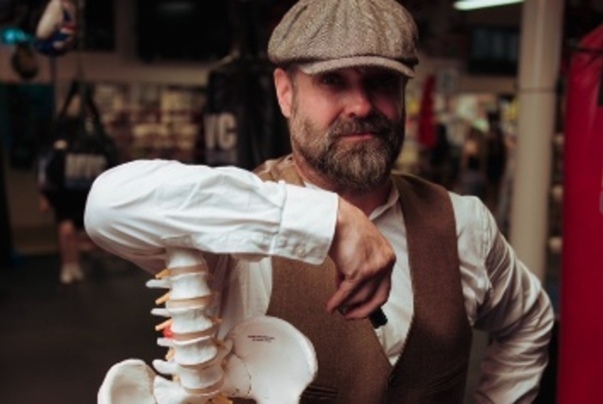 Author and storyteller Roy Maloy wearing a peaked cap and a button-down waistcoat, with his arm resting on a cast of a skeleton's spine.