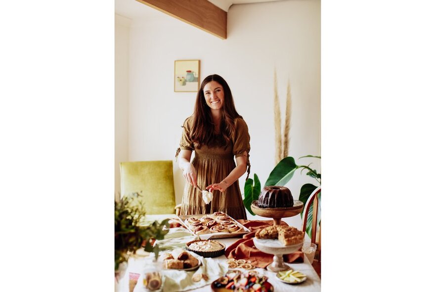 Cookbook author Georgia Irwin standing behind a table that is filled with assorted baked items on plates, in a residential dining room.