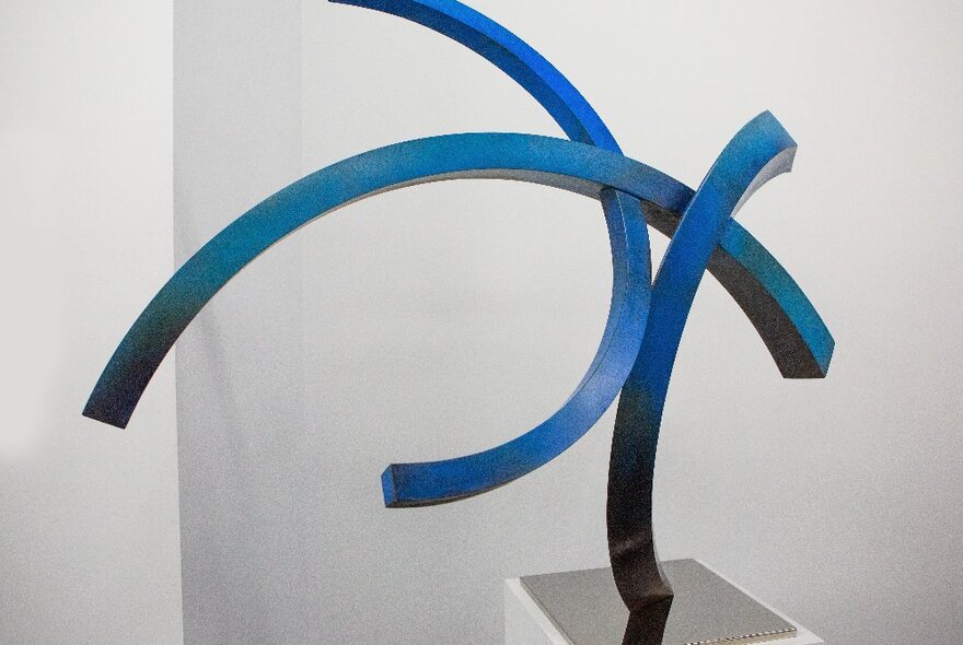 Large free standing sculpture on a plinth, comprising three intersecting and curved blue steel rods.