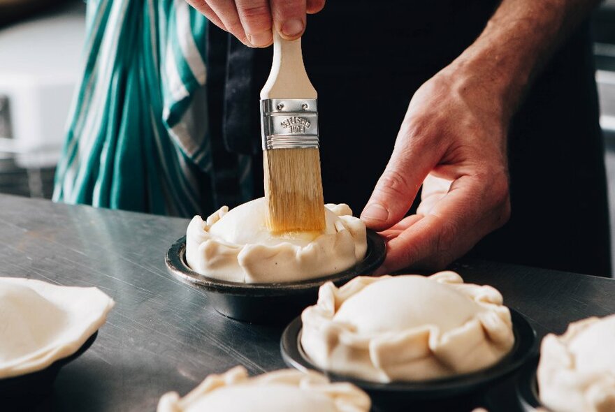 A person brushing the tops of uncooked pies with a pastry brush.