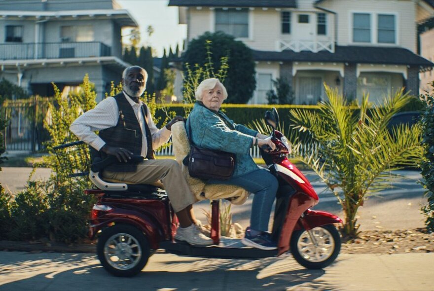 Two older people seated on a motorised scooter in front of an apartment block; a still from the film Thelma.