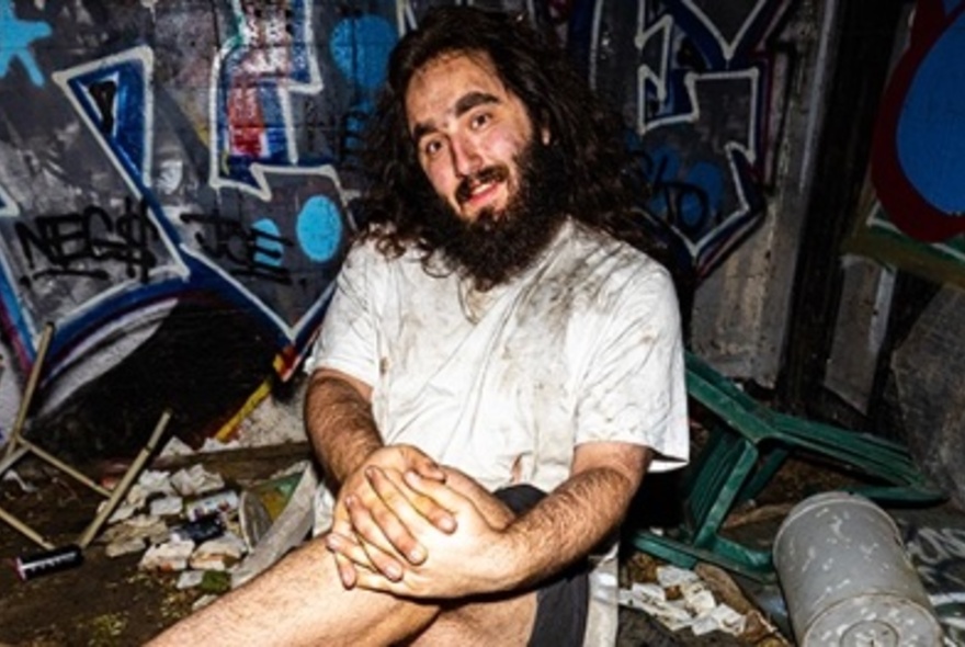 A grubby bearded man sits with his hands clasped on his crossed knee in a graffitied room. 