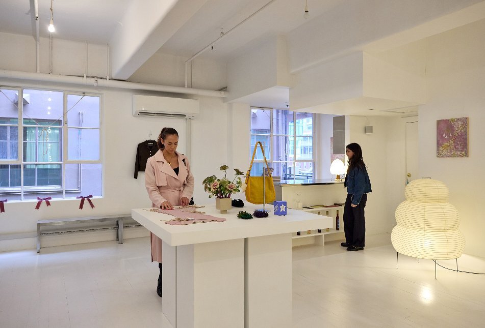 Two women are browsing items in a minimalistic clothing and homewares store with white floors, walls and ceiling.