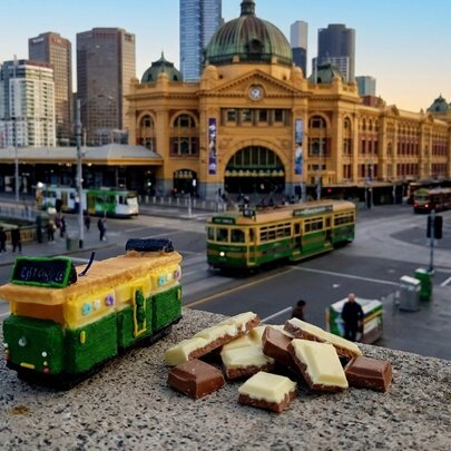A mini Melbourne tram made out of chocolate, sitting of a concrete ledge next to top deck Belgian chocolate pieces.