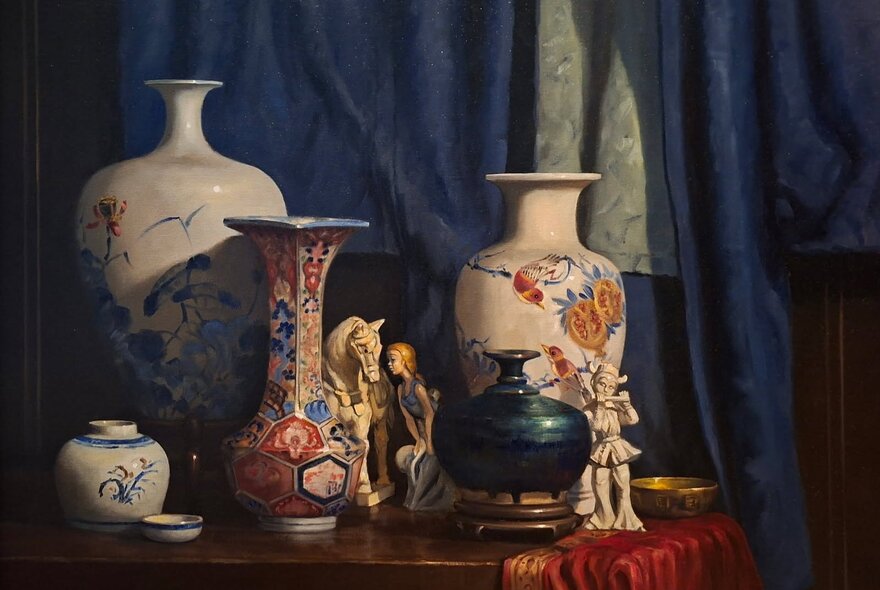 Still life oil painting of decorated porcelain vases and small objects, resting on a wooden sideboard, with a heavy blue curtain backdrop.