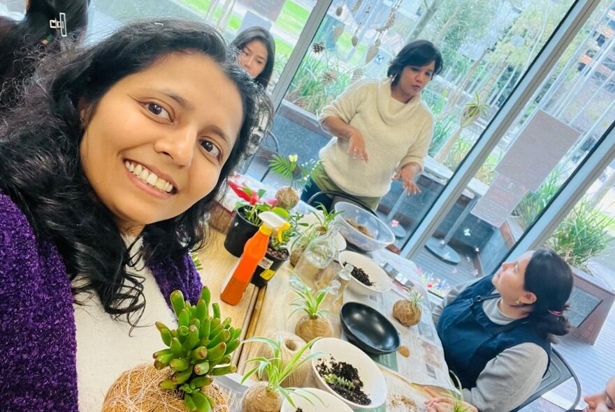 People at a workshop making kokedama balls, the work table covered in plants and soil, and full length windows behind them.