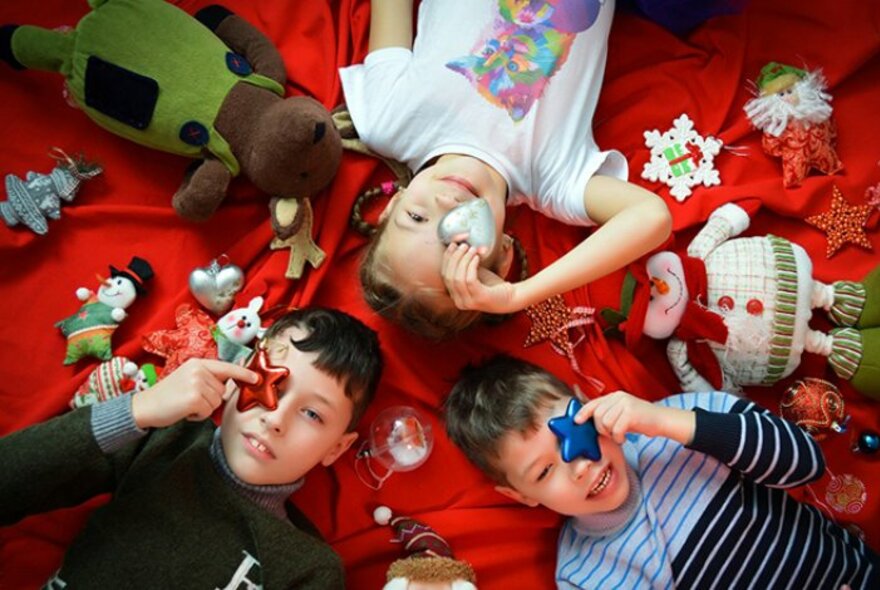 Looking down from above at three children who are lying on their backs on a red blanket, each of them holding a Christmas decoration over one of their eyes, the floor around them covered in toys and festive ornaments. 