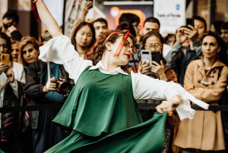 A Persian dancer wearing a traditional green top and skirt, a crowd watching as she dances.