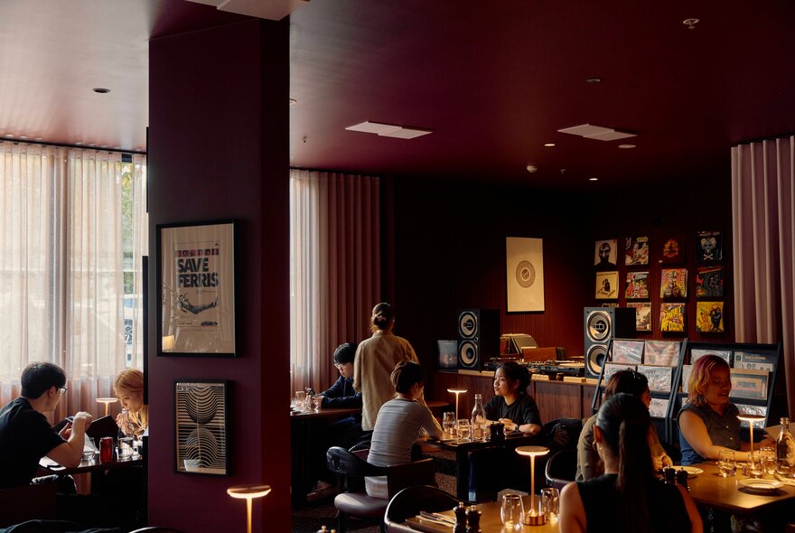 Burgundy walls and walnut-toned interior of Common Cuts restaurant, with patrons dining at tables, and a DJ vinyl record booth in the corner.