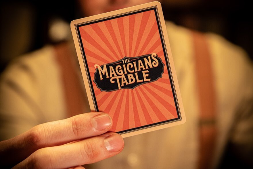 A hand holding up a custom playing card from 'The Magician's Table', an immersive close-up magic experience.