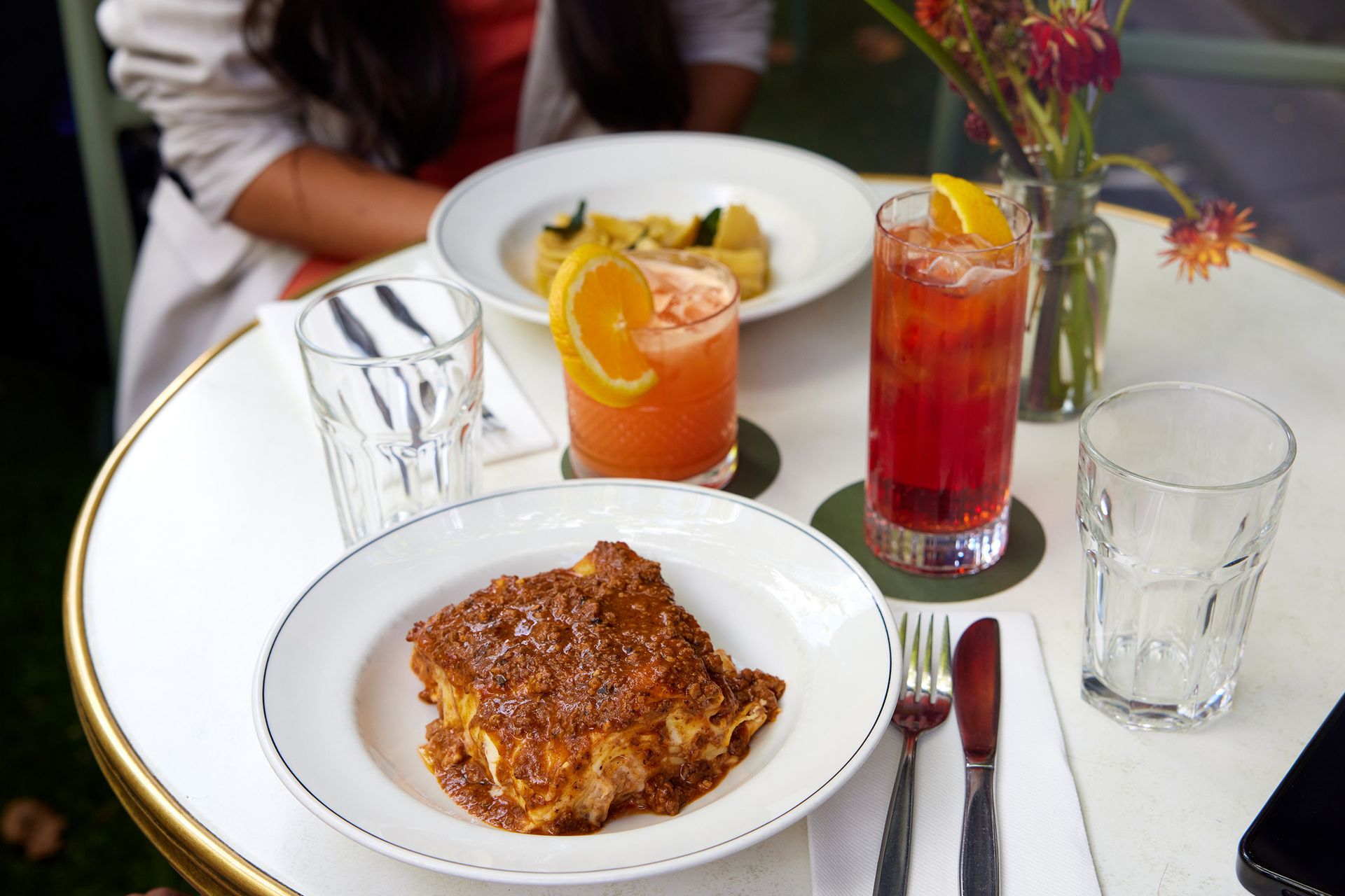 A slice of lasagne covered in meat sauce and served on a white plate. There are two cocktails and another bowl of pasta on the table.