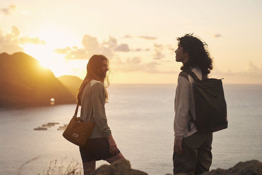 Two people standing at the top of a peak with the sun setting over the ocean in the distance, both carrying a Fjallraven bag.