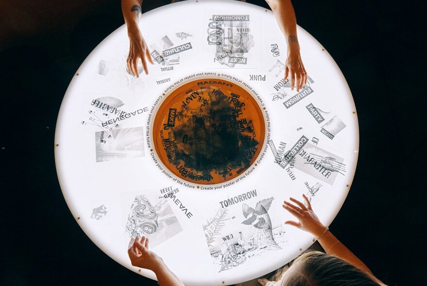 People's hands touching an illuminated circular interactive table in a museum exhibition space.