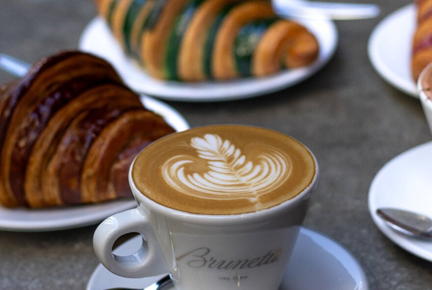 A cappuccino in a cup, with two croissants on plates in the background.
