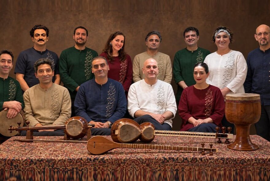 A group of Persian musicians posing in two rows in front of a table showcasing four different types of Persian instruments.