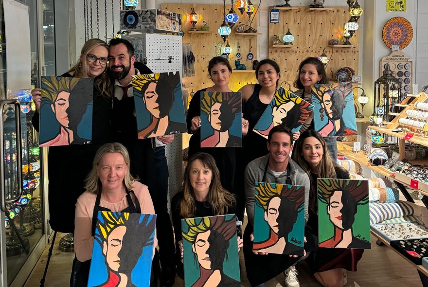 A group of people at a Paint and Sip class, holding their artworks and smiling.