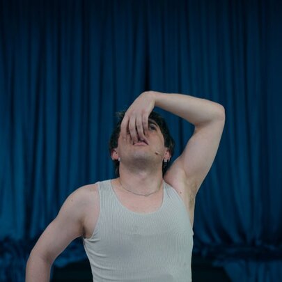 A contemporary dancer wearing a grey singlet, looking up with his hand in front of his face; a blue stage curtain behind him.