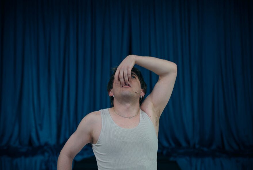 A contemporary dancer wearing a grey singlet, looking up with his hand in front of his face; a blue stage curtain behind him.