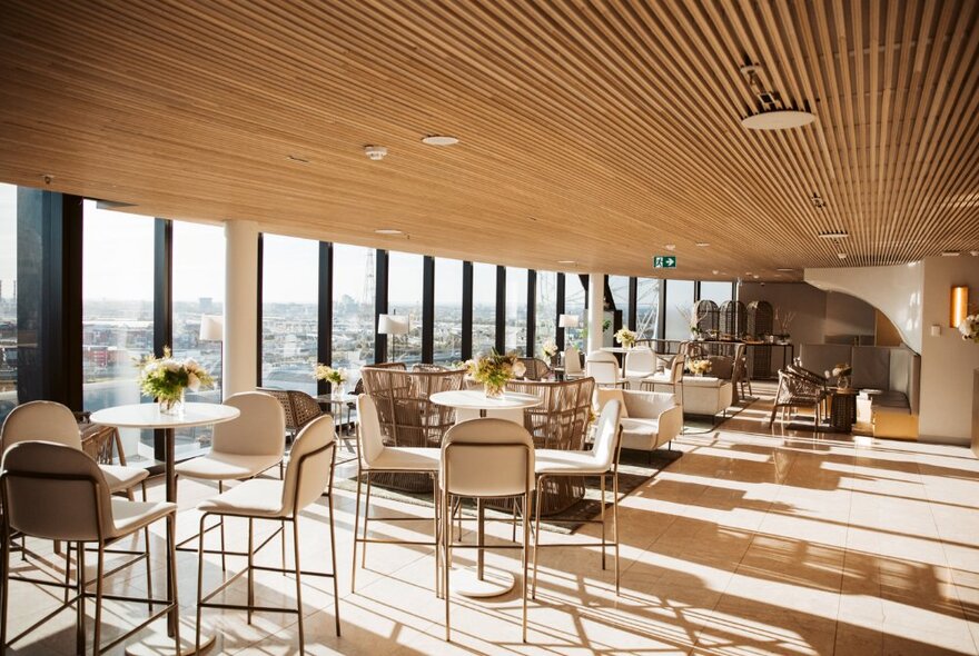 Sun-drenched penthouse lounge bar with tables and chairs, panoramic windows and striped wooden ceiling.