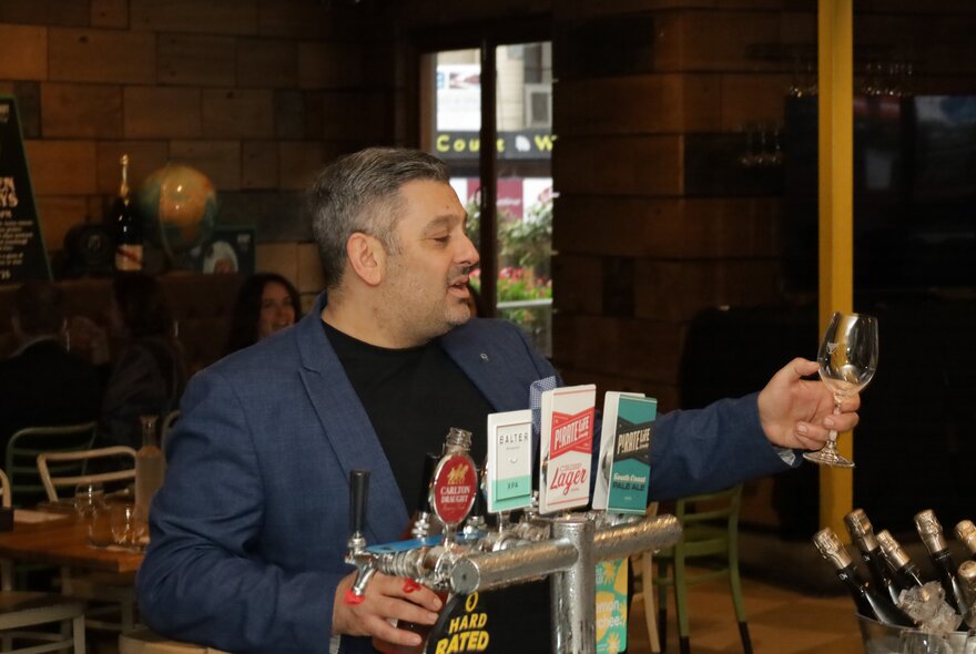 A man in a blue jacket behind a bar with beer taps, holding a wine glass in front of himself.