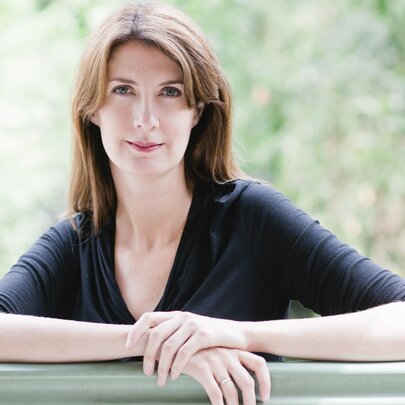 Anna Goldsworthy, a middle-aged woman wearing a black top, her arms are resting on a bench in front of her, she has shoulder length brown hair.