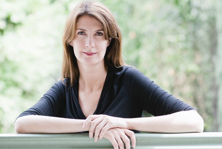 Anna Goldsworthy, a middle-aged woman wearing a black top, her arms are resting on a bench in front of her, she has shoulder length brown hair.
