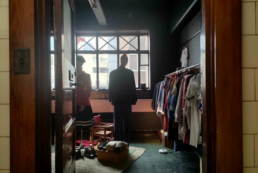 The open doorway to a small studio space that shows a rack of clothes on the right side wall, a large window against the back wall and two mannequins in the centre of the room.