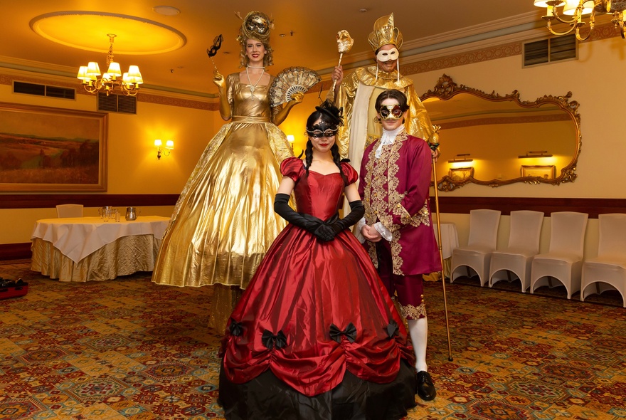 Masked women and men wearing period costumes standing in a hotel banquet room with elaborate decor.