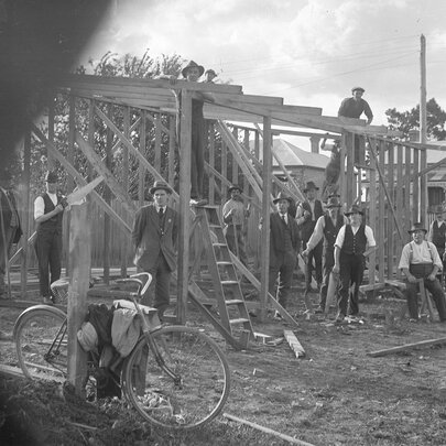 An early photograph of men in suits and hats on a building site, looking through a fence or gateway. 