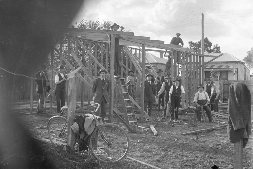 An early photograph of men in suits and hats on a building site, looking through a fence or gateway. 